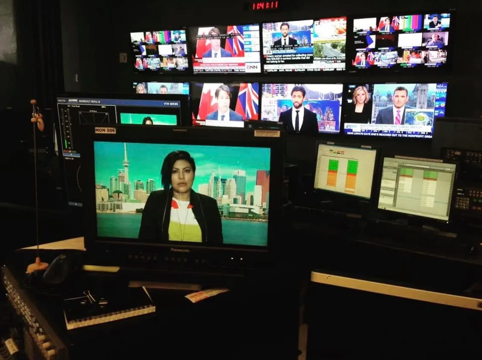 A television control room: a woman speaks on the main monitor, below a wall of news screens.