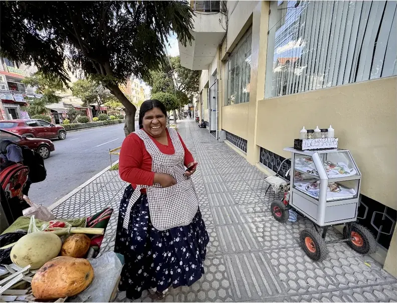 A street vendor in an apron stands beside her cart and her vegetables.