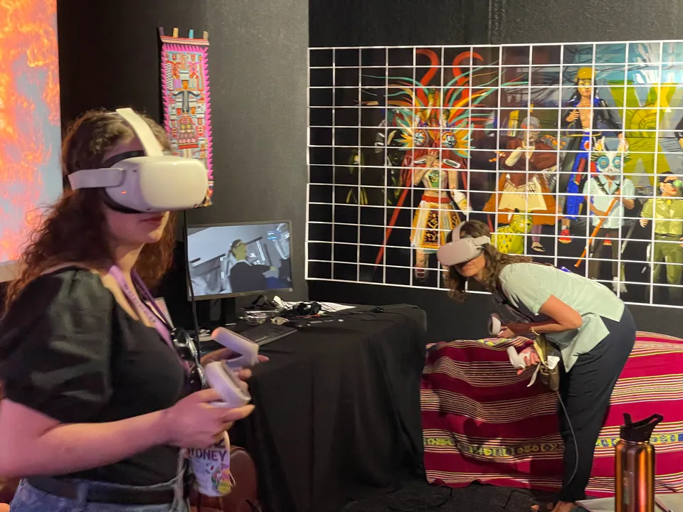 Dos visitantes con visores de realidad virtual en una mesa cubierta con un textil andino, frente a un muro con obras de Prison X.