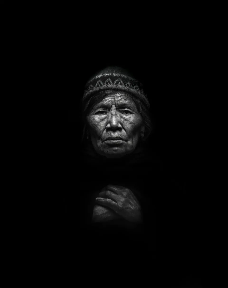 Un retrato hecho con IA, en blanco y negro: una anciana andina con gorro tejido y las manos juntas.