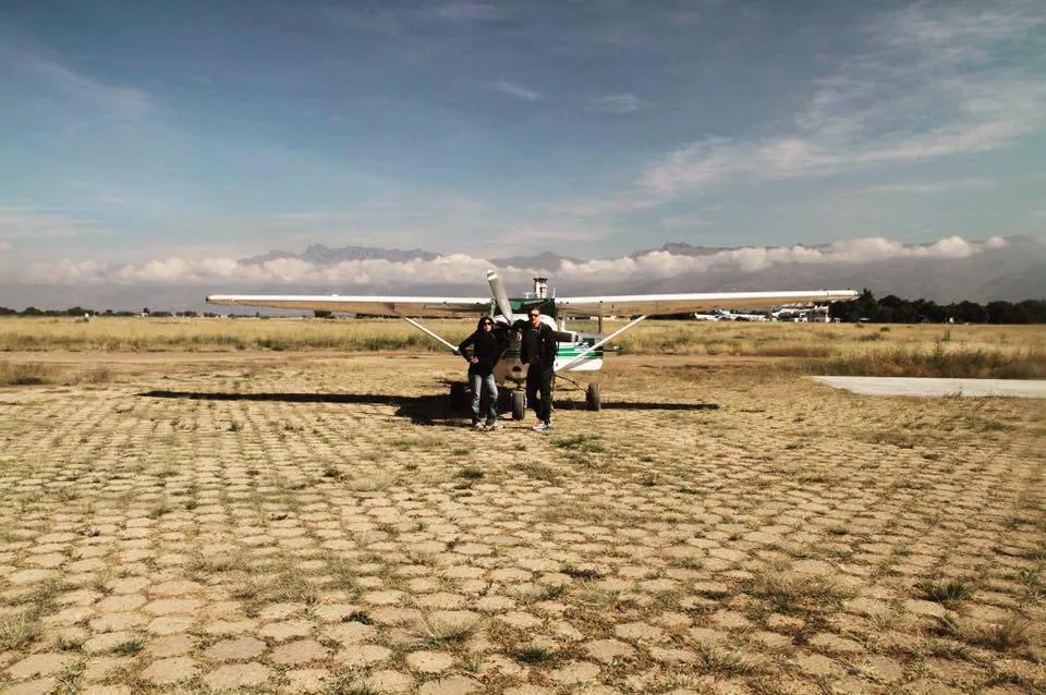 Dos personas junto a una avioneta en una pista de aterrizaje de piedra, bajo un cielo amplio.