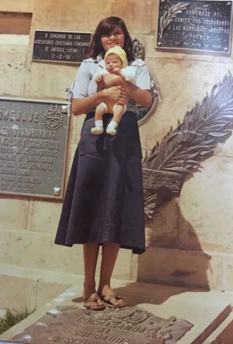 Una joven sostiene a un bebé junto a placas conmemorativas, en una fotografía antigua a color.