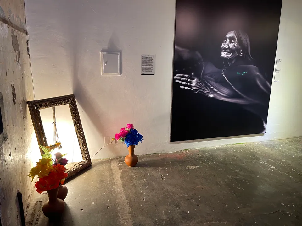 Un gran retrato de una anciana cuelga encima de un espejo y de jarrones con flores sobre un piso de cemento.