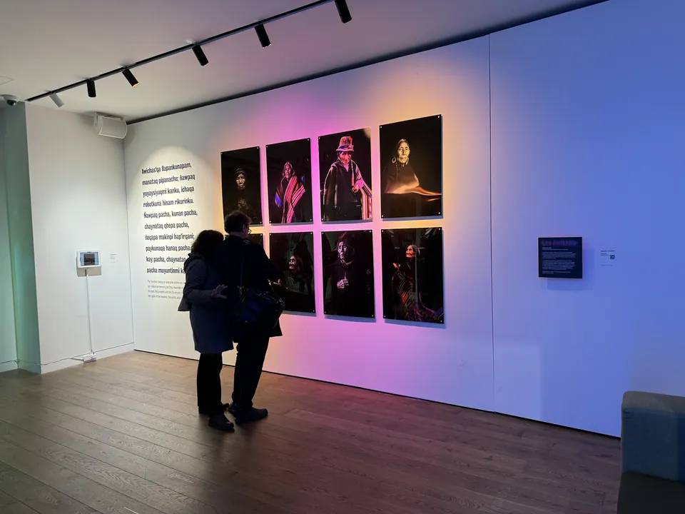 Dos visitantes están de pie frente a un muro de ocho retratos iluminado en rosado y azul.