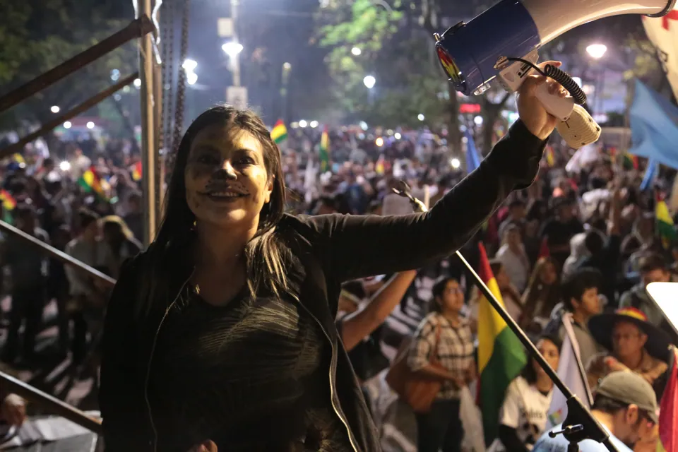 Una mujer levanta una cámara sobre una multitud de noche, con banderas bolivianas.