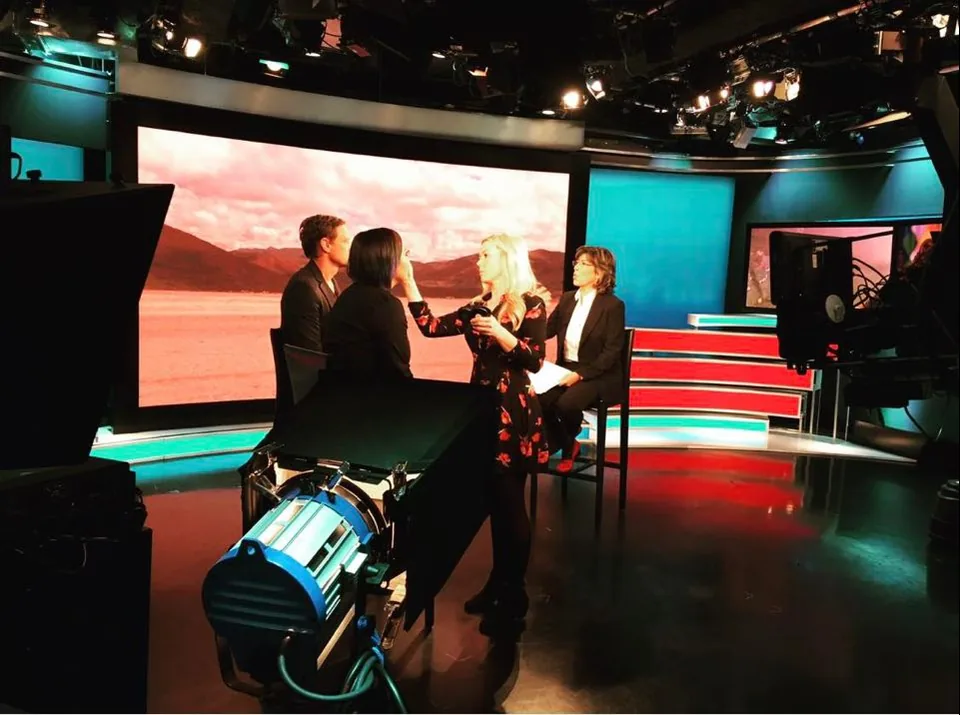 A television studio before a broadcast: a make-up artist works on a guest at the desk, beside two other people.