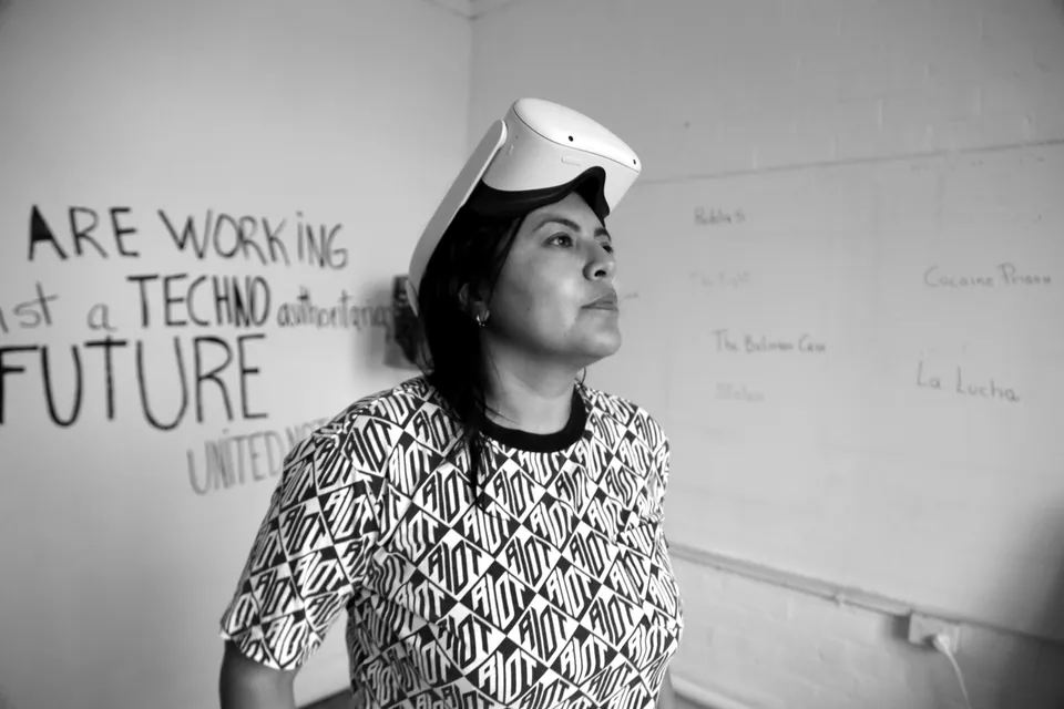Una mujer con un visor de realidad virtual en la frente, delante de un muro con las palabras: we are working against a techno authoritarian future.