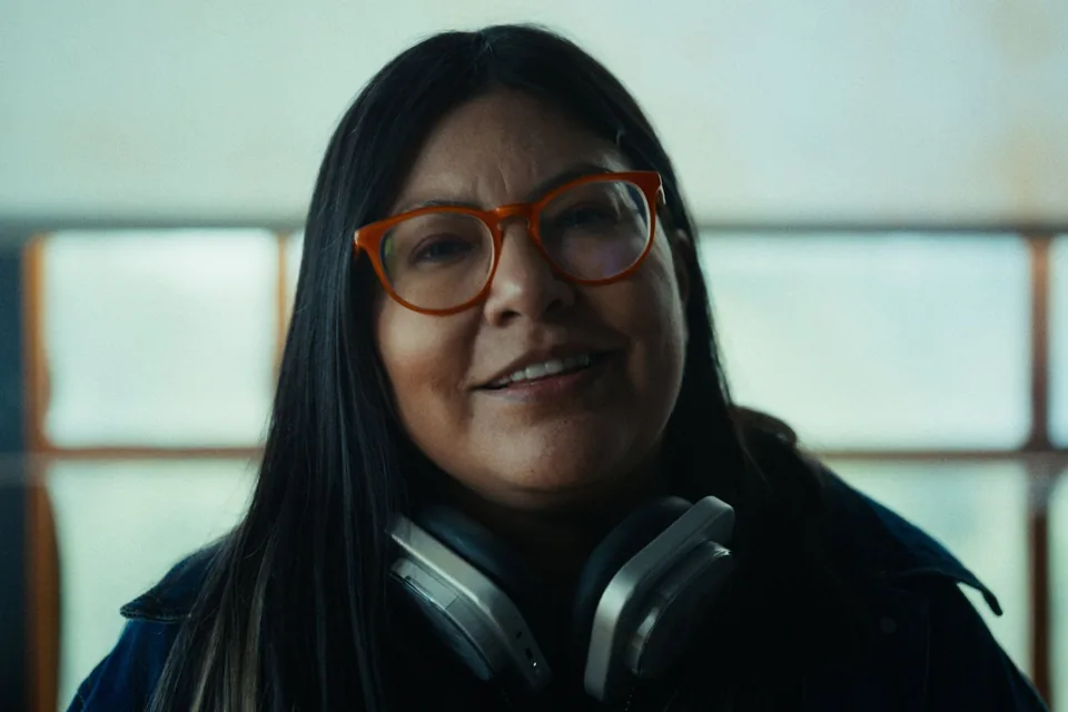 A woman with long black hair and orange glasses, headphones round her neck, smiles at the camera.