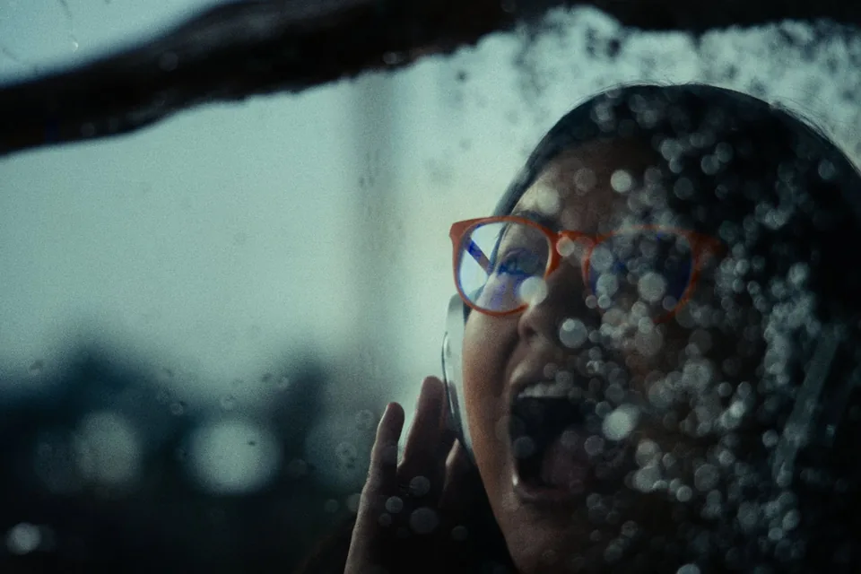Through drops of rain on the glass, a woman in orange glasses holds a hand to her headphones and calls out.