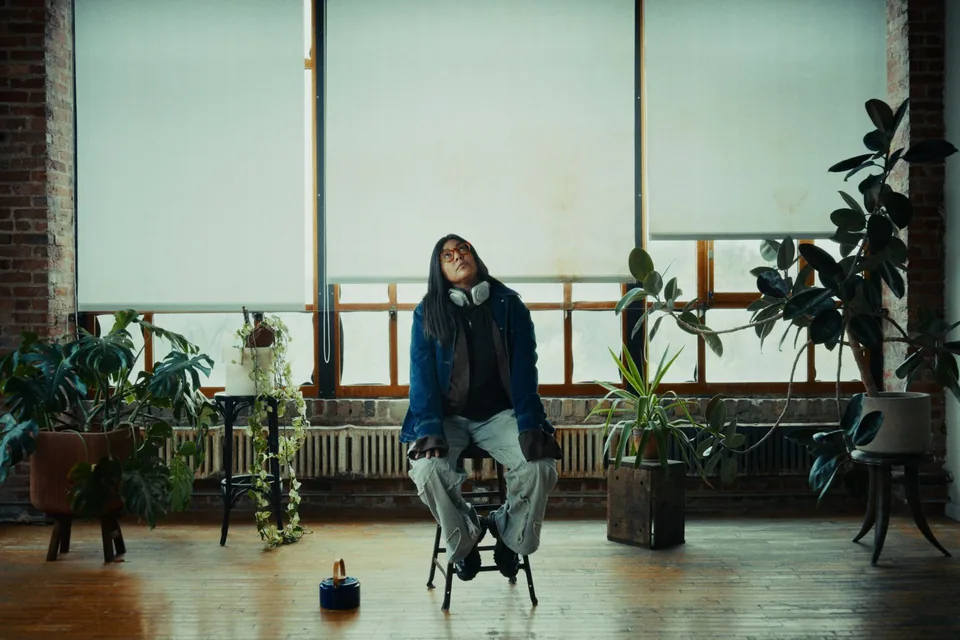In a loft of brick walls and tall windows with white blinds, a woman in a denim jacket, headphones round her neck, sits on a stool among plants and looks up.
