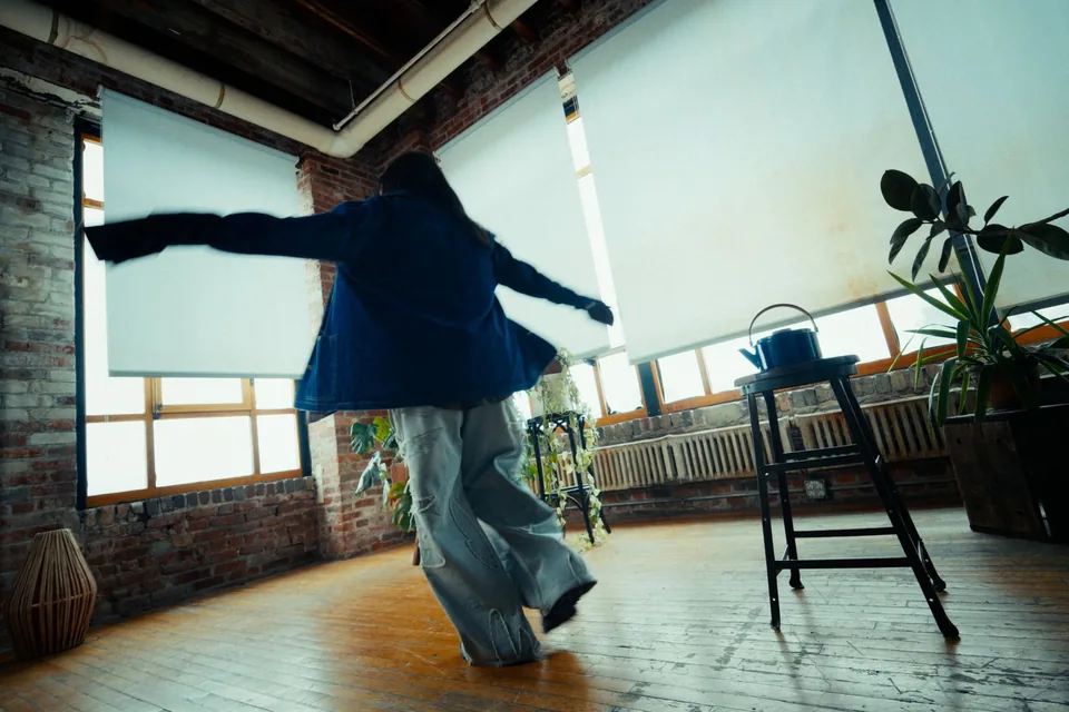 A woman in a denim jacket and wide pale trousers spins with her arms out across the wooden floor of a bright loft.