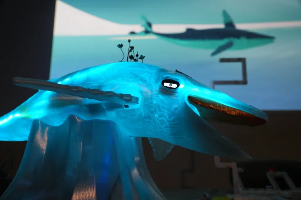 The printed whale glows blue on her stand, copper flowers on her back and a small round screen for an eye; behind her on the wall, her digital self swims.