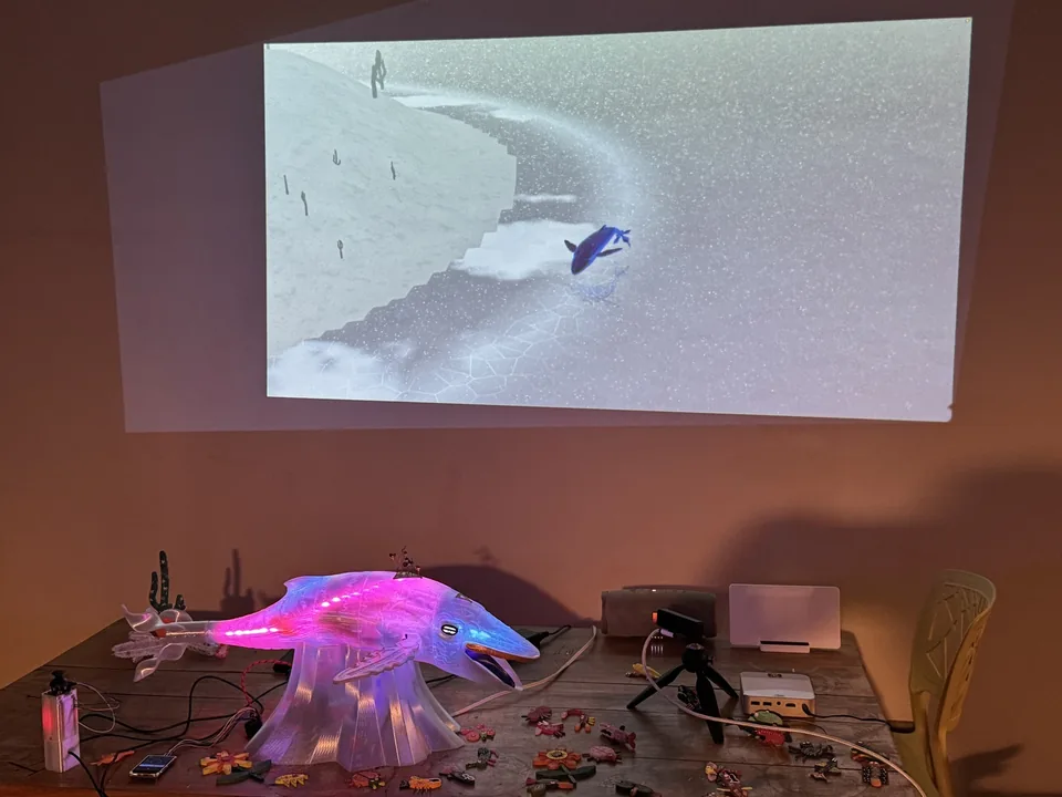 The studio in the evening: the printed whale glows magenta on a table scattered with wires and painted flowers; on the wall a projected whale dives beside a snowy island with cacti.